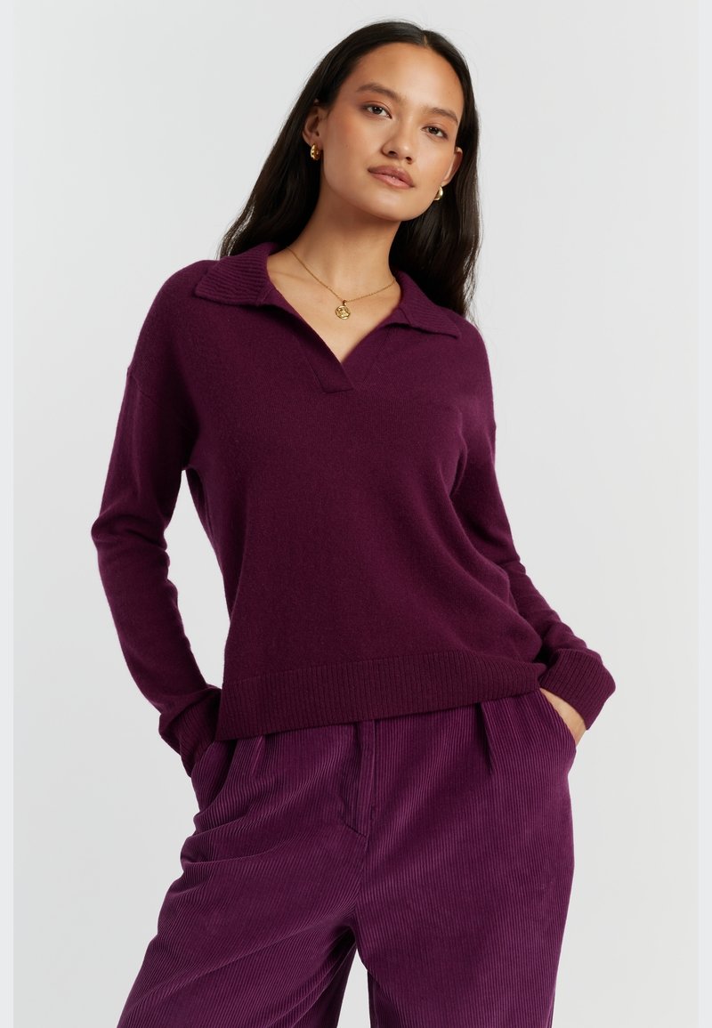 Lila gestrickter Polo-Pullover mit V-Ausschnitt, gerippten Bündchen und Saum. Kombiniert mit passenden Cordhosen, die vertikale Streifen zeigen.