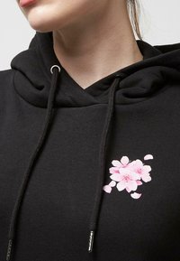 Hoodie noir avec cordon de serrage, orné d'un imprimé floral rose sur la poitrine. Construit en tissu doux avec une texture lisse.