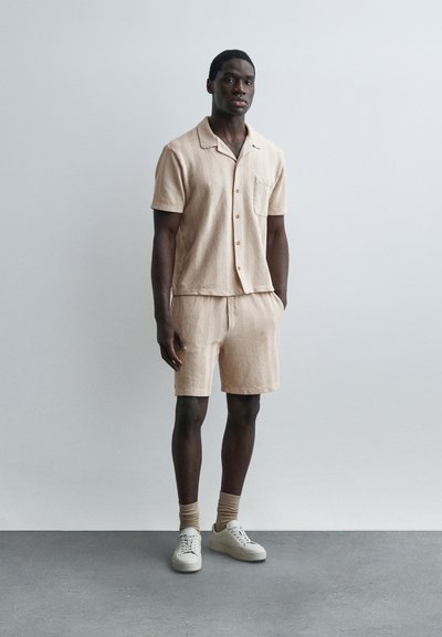 Homme debout à l'intérieur portant une chemise à manches courtes beige assortie à son short, avec des chaussettes beiges et des baskets blanches.