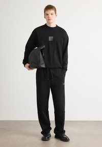 Zwarte sweatshirt met lange mouwen en een vierkant logo-label, gecombineerd met zwarte sweatpants met trekkoord. Het model houdt een zwarte tas vast.