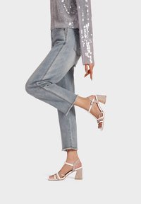 Sandali con tacco alto e cinturini di color rosa chiaro, con un tacco a blocco di un colore chiaro e texture. Indossati con jeans blu a vita alta e una maglietta grigia con paillettes.