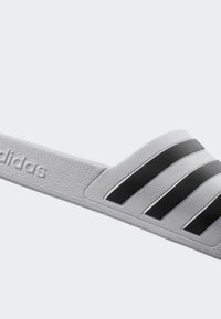 Šedé skládací sandály vyrobené z texturovaného materiálu s širokým páskem, který má tři černé pruhy a vyražené logo na boku.