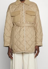 Veste matelassée beige avec un col, deux grandes poches à rabat et une ceinture à la taille. Texture lisse avec un passepoil contrastant plus clair.