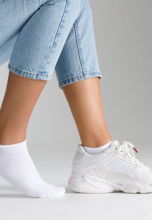 Białe sportowe sneakersy z teksturowanymi akcentami i wyściełaną podeszwą, w połączeniu z białą skarpetą do kostki, noszone z jasnoniebieskimi dżinsami o krótkiej nogawce.