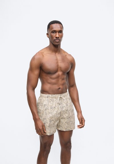 Αθλητικός άνδρας που στέκεται, φοράει μπεζ μαγιό με σχέδια, ουδέτερη έκφραση, με φόντο απλό ανοιχτό γκρι.