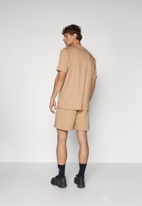 Beige short-sleeve t-shirt and matching shorts, both in a smooth fabric. Black shoes and socks complete the outfit, viewed from the back.