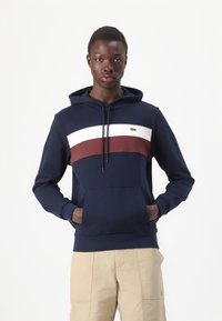 Felpa con cappuccio blu navy con una striscia bordeaux e bianca sul petto, dotata di tasca a marsupio e piccolo logo ricamato a sinistra.