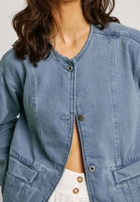 Chaqueta de mezclilla azul claro con cuello redondo, cierre de botones a presión, dos bolsillos frontales y un corte relajado con detalles de costura visibles.