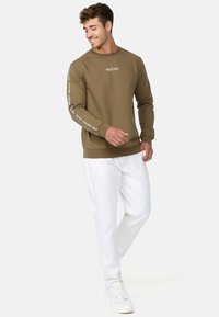 Olive green sweatshirt with long sleeves, ribbed cuffs, and white logo text on sleeves. Paired with white pants and sneakers.
