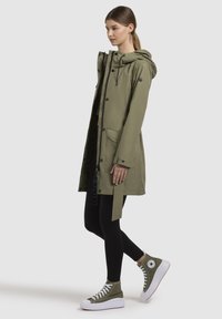 Olijfgroen waterdichte parka met een capuchon, voorzakken en een trekkoord. Draagt over zwarte leggings en groene high-top sneakers.