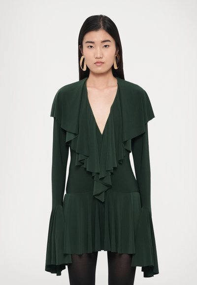 Green dress made of soft fabric, featuring layered ruffles, a deep V-neck, and flared long sleeves. Worn with large, round gold earrings.