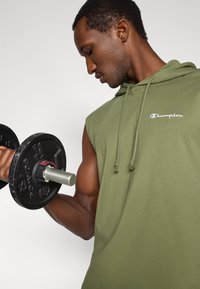 Φούτερ χωρίς μανίκια σε ελαιόχρωμο με κορδόνι, με το λογότυπο "Champion". Ο μυϊκός τόνος είναι ορατός ενώ κρατά ένα μαύρο αλτήρα.