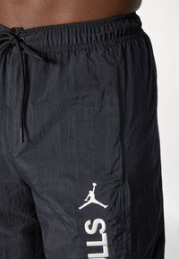 Black shorts made of lightweight material with an elastic waistband. Features a white logo and text print on the side. Textured fabric.