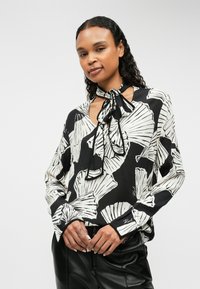 Blusa nera con stampa di foglie bianche, dotata di colletto a fiocco, maniche lunghe e vestibilità rilassata. La texture simile al satin conferisce morbidezza.