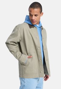 Ανοιχτό πράσινο βαμβακερό μπουφάν με καφέ γιακά, κλείσιμο με κουμπιά και δύο εμπρόσθιες τσέπες, φορεμένο πάνω από μπλε φούτερ με κουκούλα και ανοιχτό μπλε τζιν.