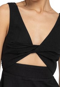 Kvinna iklädd en ärmlös svart klänning med en vriden framsida och ett urklipp under bysten, matchad med ett delikat guldchain-halsband.