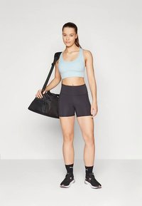 Modro-svetel športni modrček, črne kratke hlače z visokim pasom in črne superge. Model nosi črno torbo čez ramo.