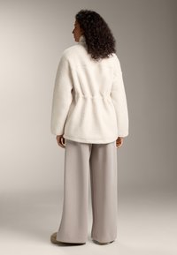 Fehér sherpa pulóver magas gallérral, elasztikus derékrésszel és bővebb fazonnal. Világosszürke, bő szárú nadrággal és bézs cipővel párosítva.