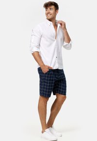 Camisa de lino blanca de botones con mangas enrolladas, combinada con pantalones cortos a cuadros en azul marino y zapatillas blancas. Ajuste casual y relajado, apto para el clima cálido.