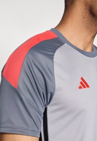  Γκρι αθλητική μπλούζα με κόκκινες λεπτομέρειες στον ώμο, κατασκευασμένη από απαλό, ελαφρύ ύφασμα. Διαθέτει μικρό κόκκινο λογότυπο στο στήθος.