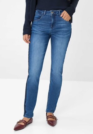Frau in blauen Jeans mit dunklen Seitenstreifen, marineblauem Pullover und burgunderfarbenen, spitz zulaufenden Flats mit Schnallenriemen, steht vor einem weißen Hintergrund.