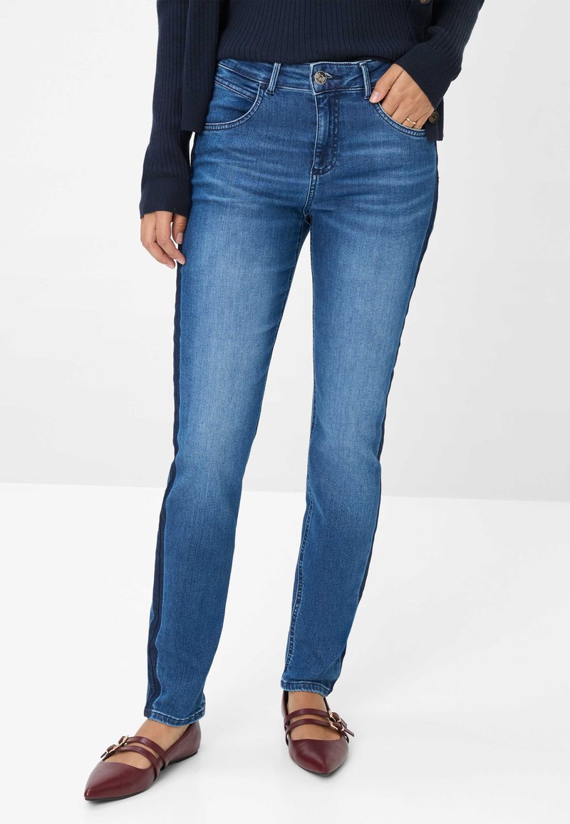 Donna che indossa jeans blu con strisce laterali scure, un maglione navy e delle ballerine bordeaux con cinturini a fibbia, in piedi su uno sfondo bianco.