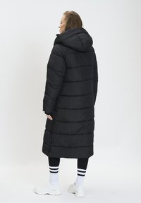 Abrigo largo negro tipo plumas con capucha, que presenta un diseño acolchonado, costuras horizontales prominentes y un corte holgado. Llevado sobre leggings negros y zapatillas blancas.