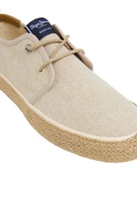 Sneaker in tela beige con soletta in juta intrecciata, punta rotonda e lacci beige. Presenta un'etichetta blu sulla linguetta e occhielli circolari.