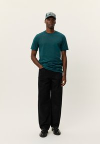 Τηλεφωνικό μπλουζάκι από βαμβάκι σε τυρκουάζ χρώμα συνδυασμένο με μαύρο παντελόνι φαρδιάς γραμμής με μια υποτονική υφή, φορεμένο με μαύρα παπούτσια και καπέλο με σχέδια.