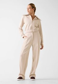 Veste beige à fermeture éclair avec un col montant, associée à un pantalon beige à jambes larges avec des plis à l'avant. Le modèle porte des chaussures marron.