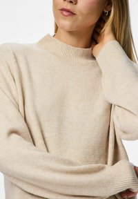 Žena s dugom plavom kosom koja nosi bež pleteni pulover, dodiruje si kosu blizu uha desnom rukom.