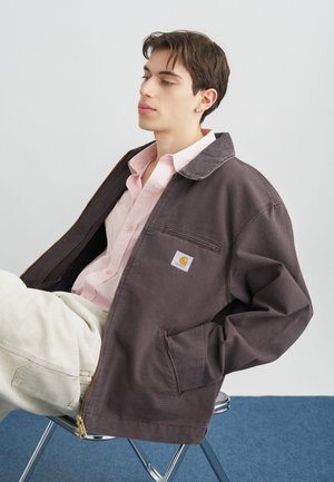 Giovane seduto su una sedia di metallo, indossa una giacca Carhartt marrone sopra una camicia rosa e pantaloni chiari, su uno sfondo semplice.