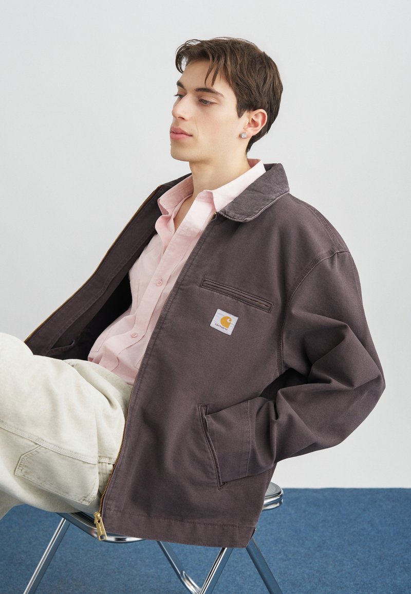 Joven sentado en una silla de metal, vistiendo una chaqueta marrón de Carhartt sobre una camisa rosa y pantalones de color claro, con un fondo liso.