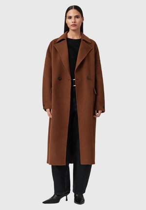 Cappotto di lana marrone con una vestibilità lunga e rilassata, ampi rever, doppi bottoni e tasche frontali, abbinato a pantaloni neri e scarpe nere a punta.