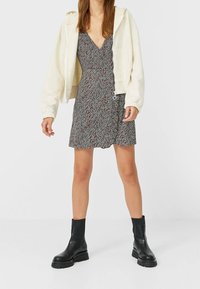 Sudadera cremosa recortada sobre un vestido negro y rojo con estampado, combinada con botas negras hasta el tobillo con suela gruesa. Texturas de tela suaves.