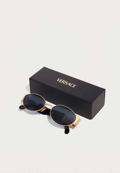 Στρογγυλά γυαλιά ηλίου με μαύρους φακούς και χρυσά πλαίσια, τοποθετημένα δίπλα σε έναν μαύρο ορθογώνιο κουτί με την επιγραφή "VERSACE" σε χρυσά γράμματα.