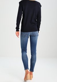 Mujer de pie de espaldas, vistiendo un suéter azul oscuro, jeans ajustados azules y zapatos de tacón beige con tacones de bloque de madera.