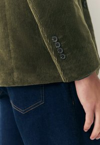 Persona vistiendo un blazer de pana verde oliva con cuatro botones en las mangas y jeans de mezclilla azul oscuro, con la mano relajada al costado.