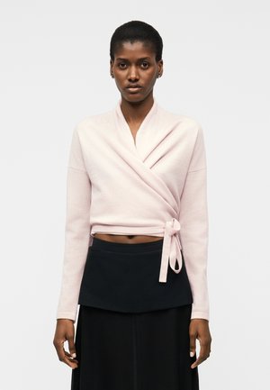 Moteris vilki šviesiai rožinį susiaučiamą megztinį, surištą ties juosmeniu, dėvi juodą sijoną, stovi prie paprasto balto fono.