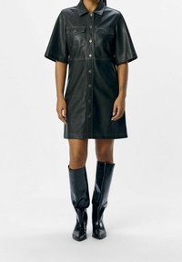 Robe chemise en simili cuir noir avec des manches courtes, deux poches avant et des fermetures à boutons. Associée à des bottes noires montantes.