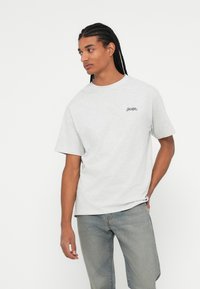 Tricou din bumbac gri deschis, cu mâneci scurte și guler rotund, având text albastru marin "JACKER" în zona stângă a pieptului. Textură netedă, croială lejeră.