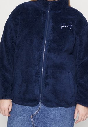 Personne portant une veste zippée duveteuse bleu foncé avec un col montant et un jean denim bleu clair.