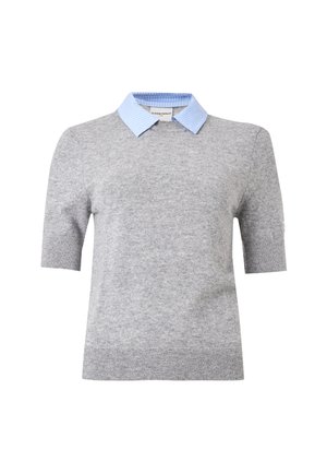 Kvinde iført en grå kortærmet sweater over en lyseblå skjorte med krave, et langt mørkeblåt skørt og sorte flade sko, stående mod en hvid baggrund.