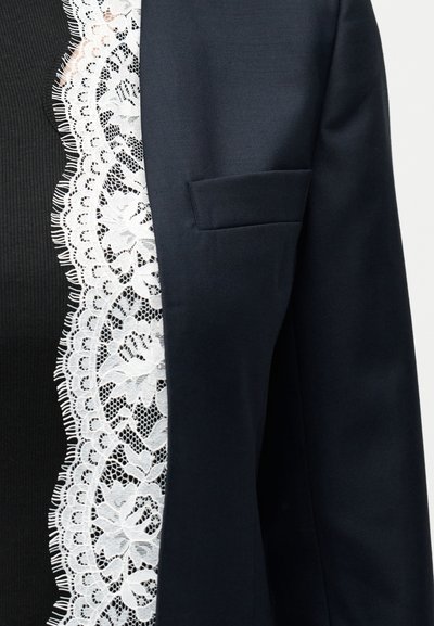 Μαύρο μπλέιζερ με λευκή φλοράλ δαντέλα πάνω από μαύρο ριμπ τοπ, εστιασμένο στην περιοχή του στήθους και των ώμων.