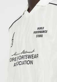 Λευκή αθλητική φανέλα με διακριτό υφασμάτινο σχέδιο, μαύρη επένδυση και τυπωμένα λογότυπα: "World Performance Studio" και "Core Sportswear Association".