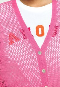Rosa stickad kofta med en lös meshdesign, prydd med silverknappar. Under finns en vit skjorta med färgglada bokstäver A, O och N.
