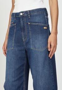 Dunkelblaue Denim-Jeans mit hoher Taille, fünf Taschen, genähten Details und einem metallischen Knopfverschluss. Markenschild auf der Rückseite.