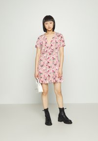 Vestido floral em rosa com mangas curtas e decote em V, apresentando uma bainha com babados, combinado com botas pretas de atacador e uma pequena bolsa branca.