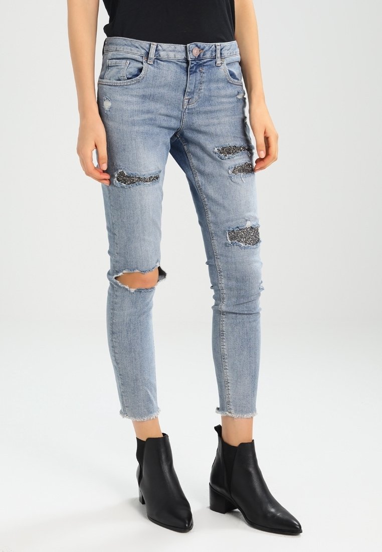 Jean skinny bleu clair déchiré avec des patchs de sequins noirs, porté avec des bottines noires et un haut noir rentré.