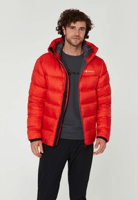 Rode pufferjack met horizontaal geborduurd ontwerp, verstelbare capuchon, rits aan de voorkant en zwarte accenten. Gedragen over een grijs shirt en zwarte broek.